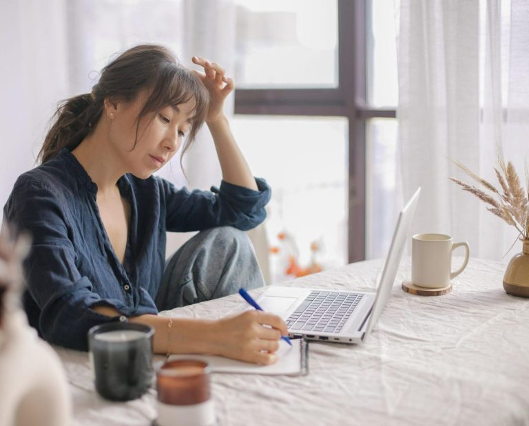 Femme assise à une table, prenant des notes dans un carnet à côté de son ordinateur portable.